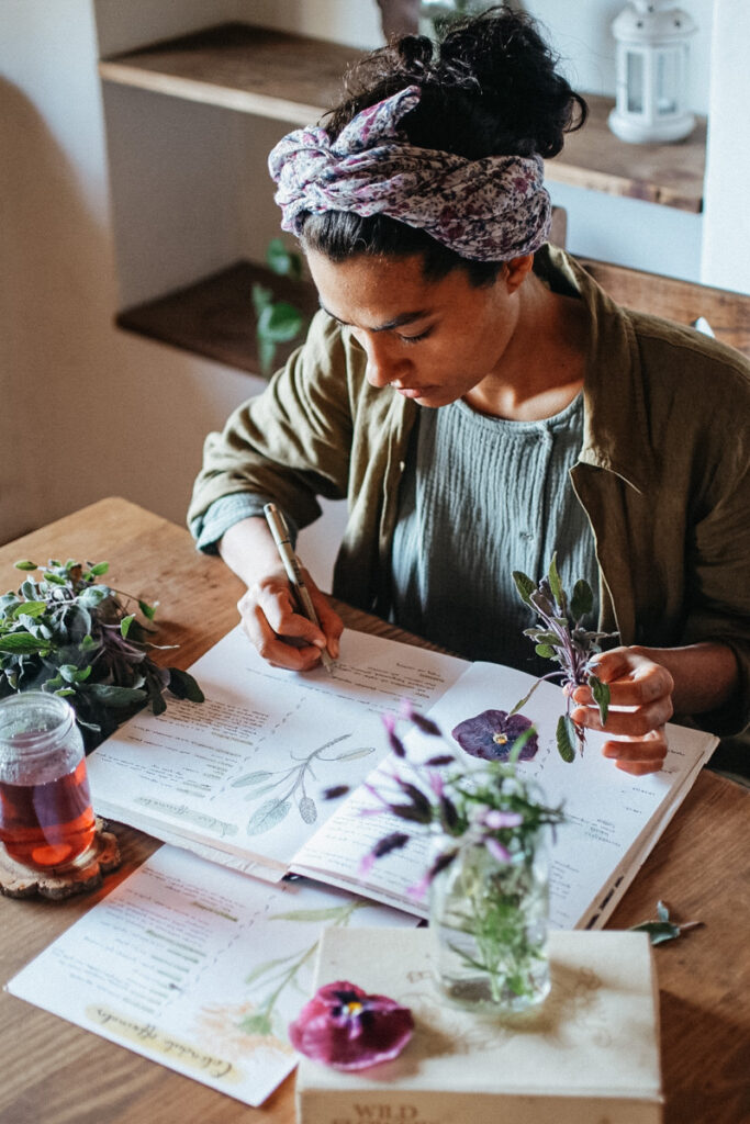 Woman with floral headwrap writes in a notebook at a wooden table, surrounded by herbs and plant specimens in a cozy workspace.