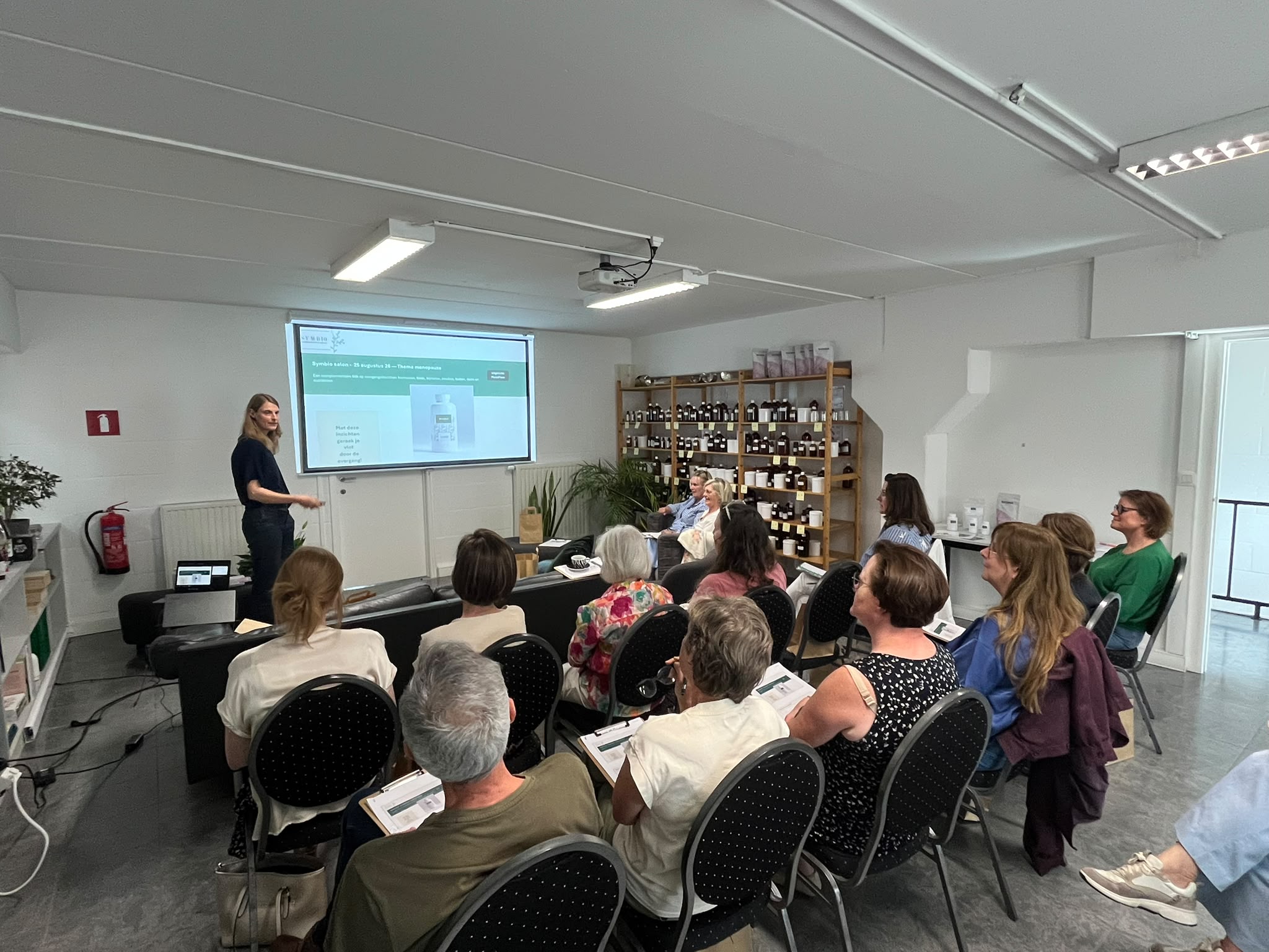Presenter stands at front of a bright meeting room giving a talk to a seated audience with a projection screen displaying slides behind her.