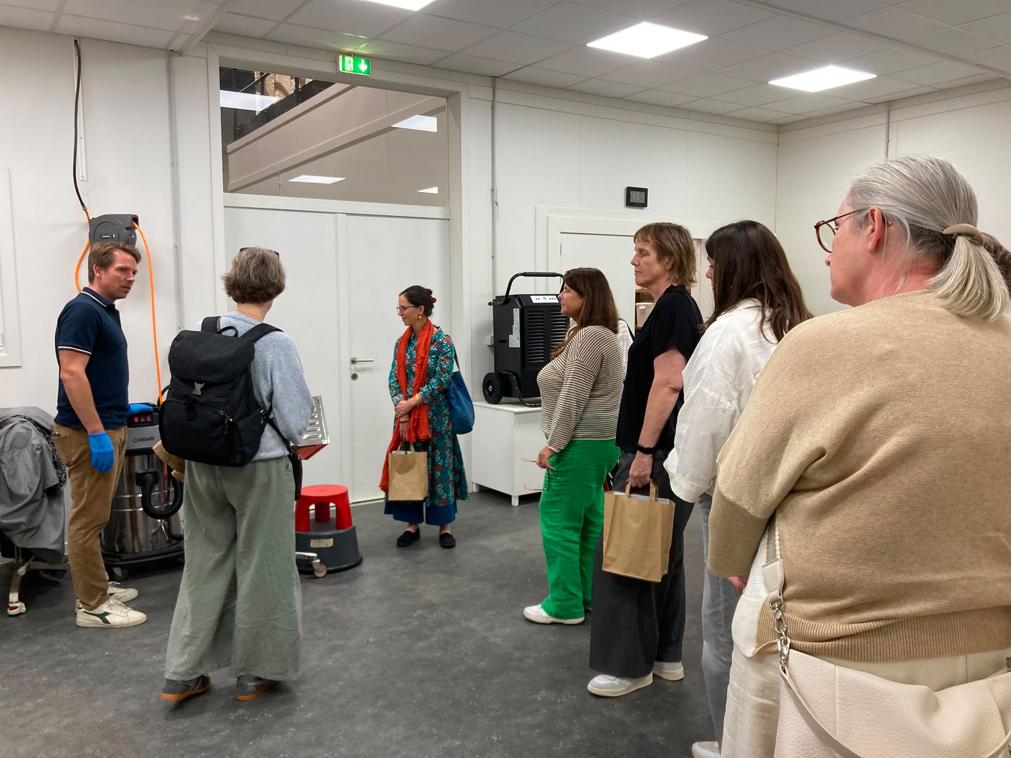 Group of adults in a workshop listening to a demonstration near industrial equipment in a white, well-lit room.