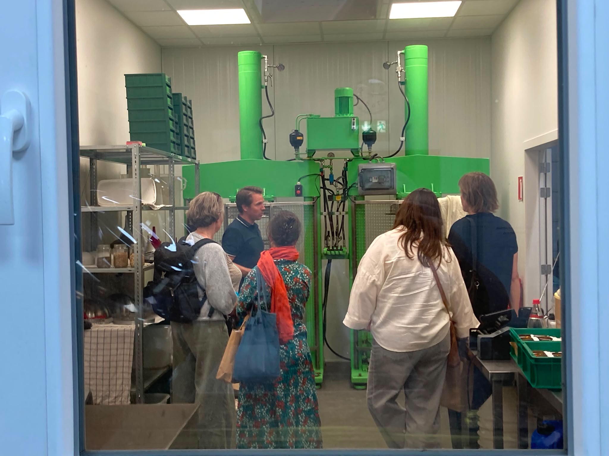 Group of people on a tour inside a bright industrial workshop, standing near large green machines and controls behind a glass partition.
